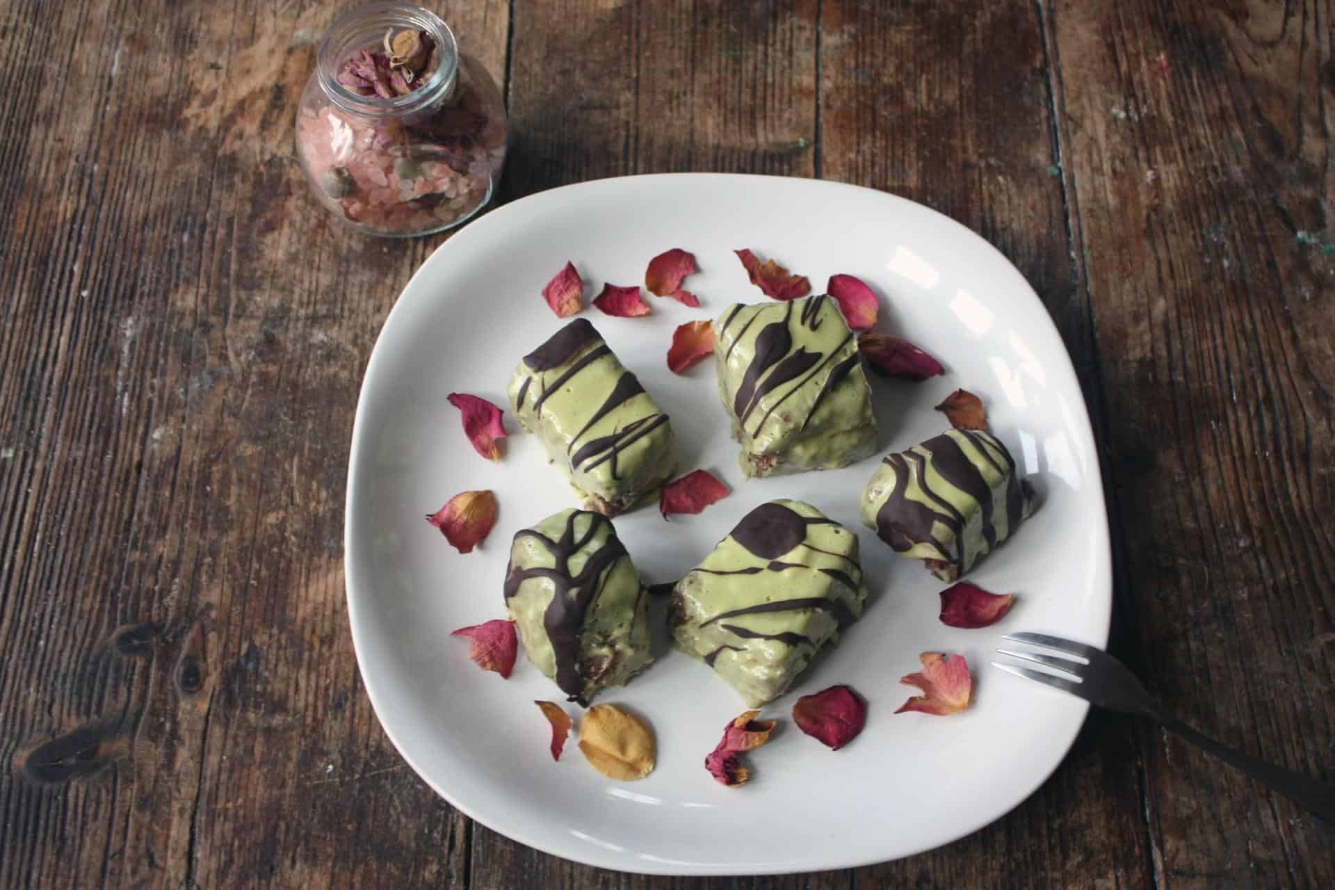 Plate containing five fondant fancies with green icing and drizzled in chocolate, sprinkled with rose petals