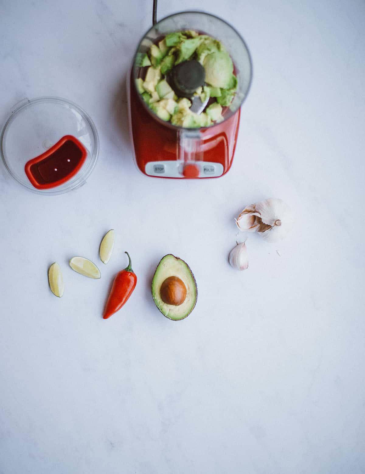 Red food processor holding avocado chunks with half an avocado, chili and garlic next to it