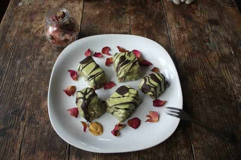 Plate containing five fondant fancies with green icing and drizzled in chocolate, sprinkled with rose petals