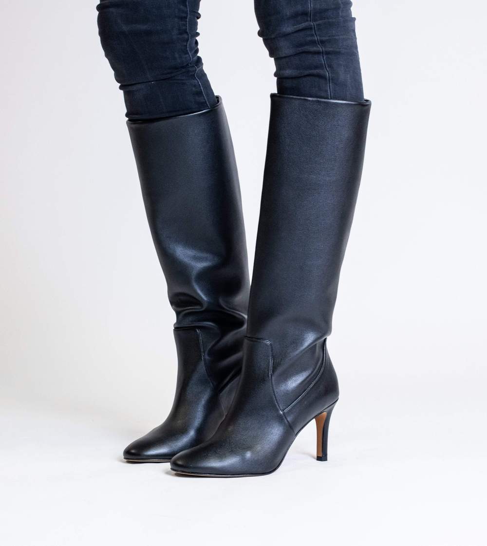 Model's legs are visible, wearing stiletto heeled black vegan leather knee high boots and dark jeans
