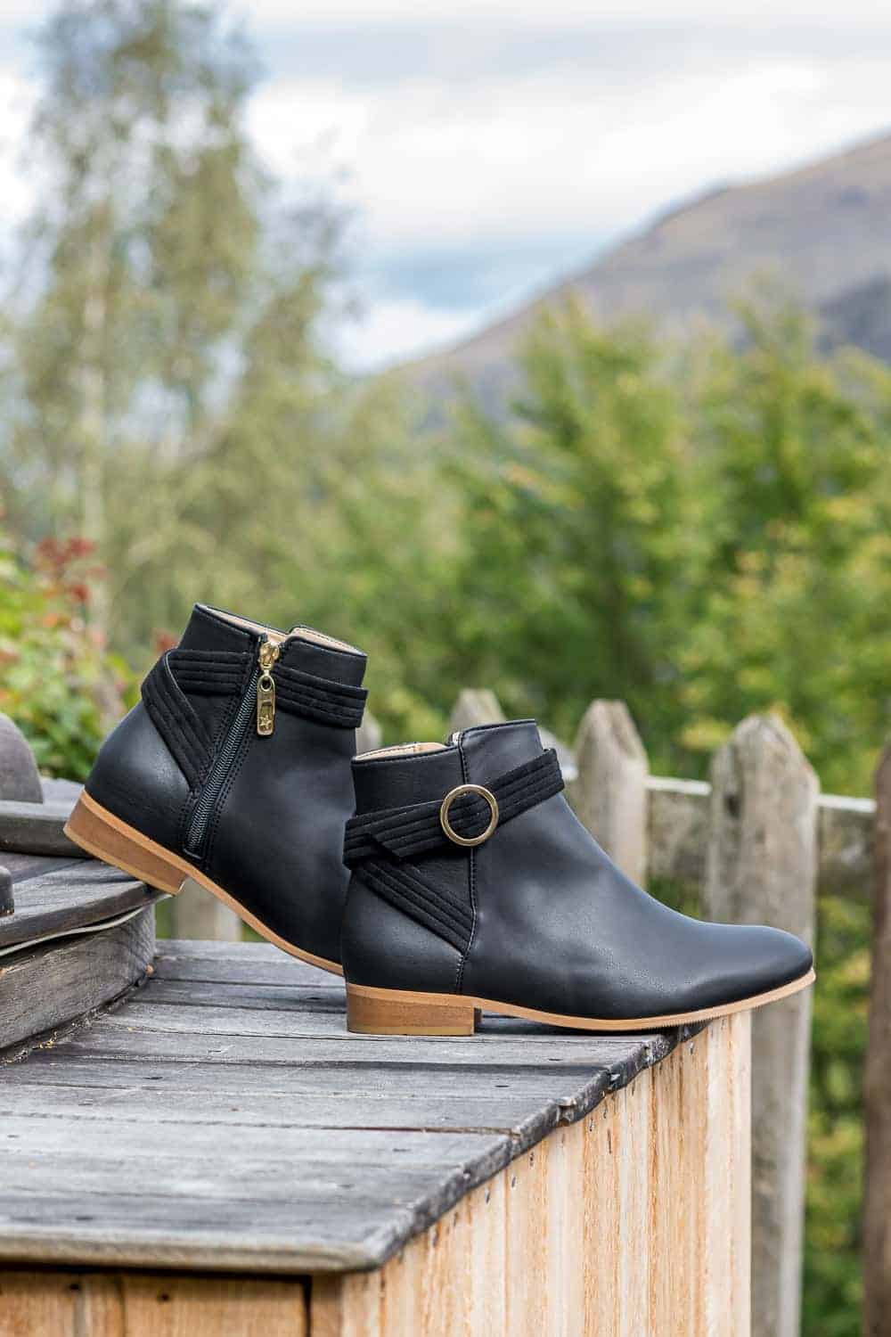 Flat black vegan leather ankle boots shown with decorative strap and round gold tone buckle on the side