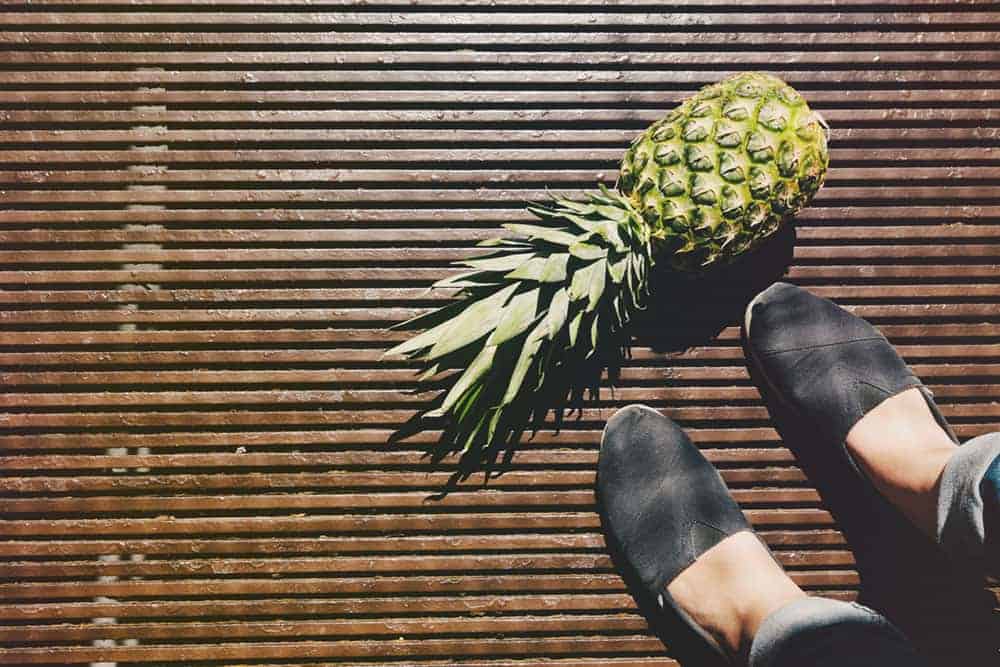 Feet wearing black shoes standing on surface made on wooden slats next to a pineapple