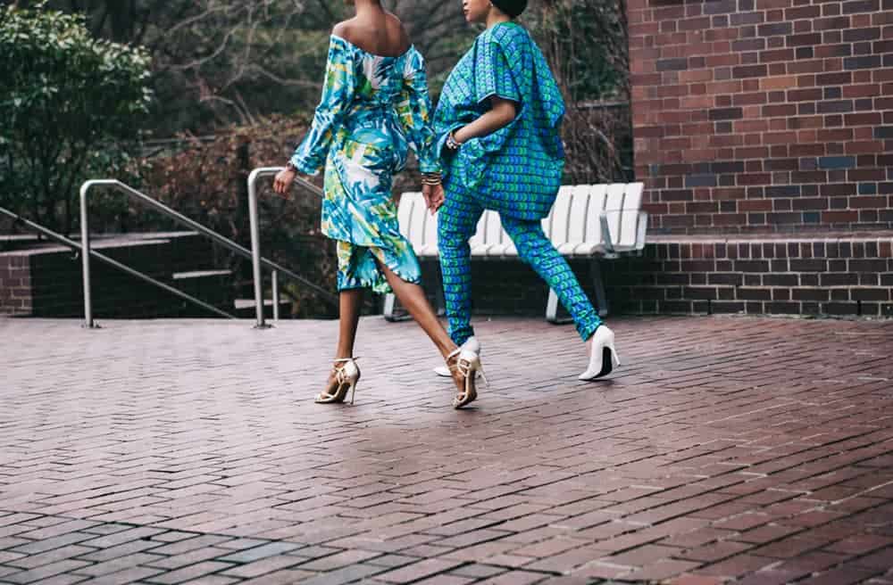 Two people in colourful print clothing shown walking on brick pathway in stiletto heels