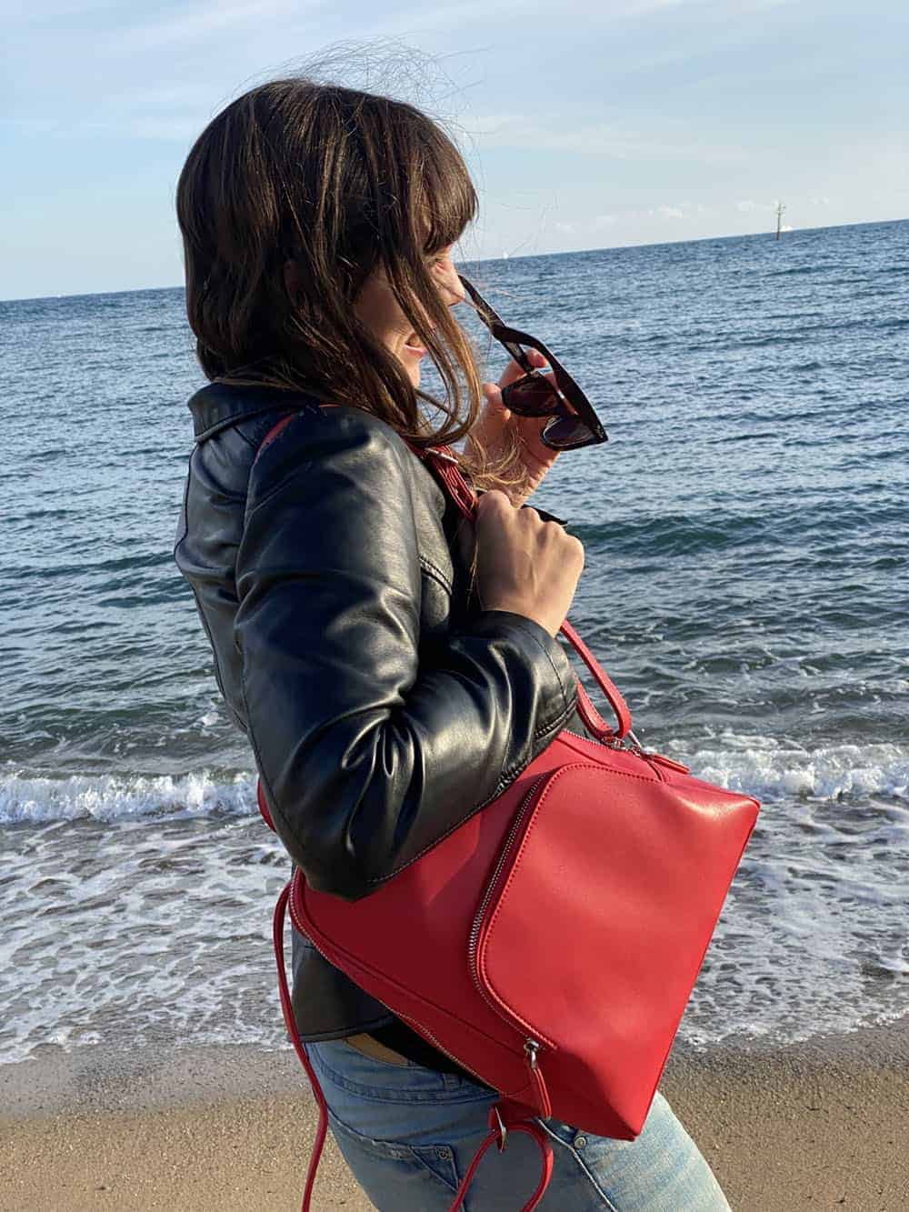 I'm holding the red vegan leather backpack from Doshi on the beach in front of the sea