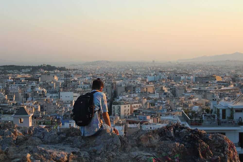 Person, viewed from behind, wearing black backpack, sitting on a rocky outcrop overlooking a city