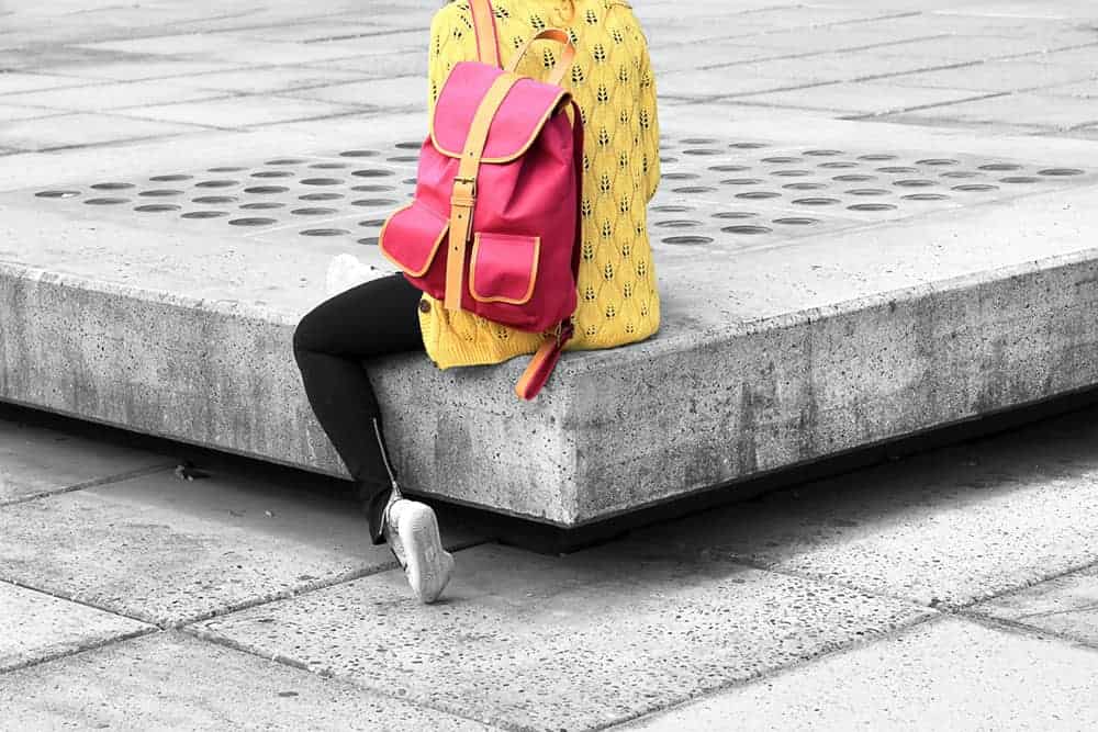 Person is shown from behind with red backpack over one shoulder. They are sitting down and wearing a yellow top and black trousers.