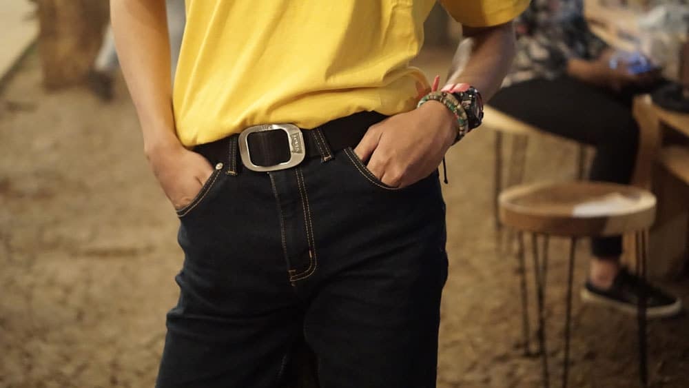 Person wearing white shirt and black jeans with a black belt stands with their hands in their pocket (only waist visible)
