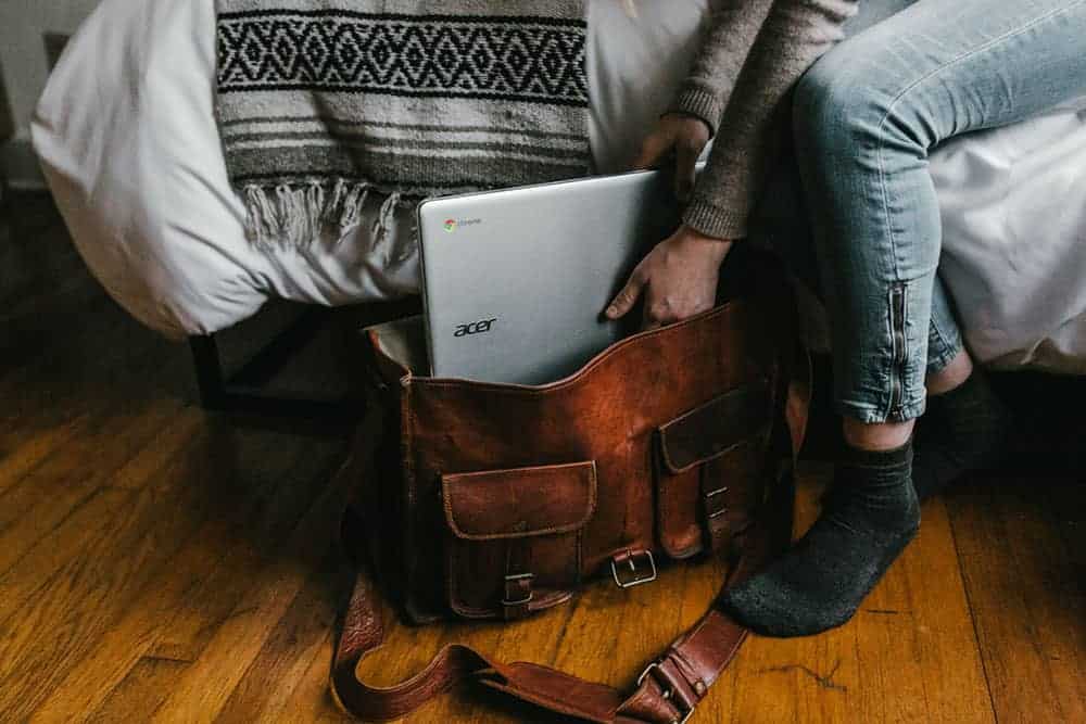 Person is shown sitting on the edge of a bed, putting a laptop into a bag