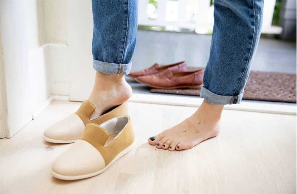 Person shown (knees down) wearing jeans and slipping their fit into pair of vegan brown suede and cream vegan leather slippers