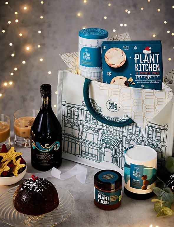 M&S bag and spread showing cookies, Christmas pudding, mince pie, chocolate hazelnut spread and liqueur sitting next to glasses containing it, with fairy lights in background