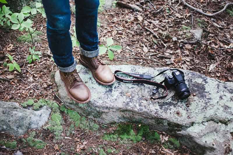 Mens vegan boots: person standing on rock wearing brown boots and jeans (only legs and feet seen). Camera is on the rock next to them.