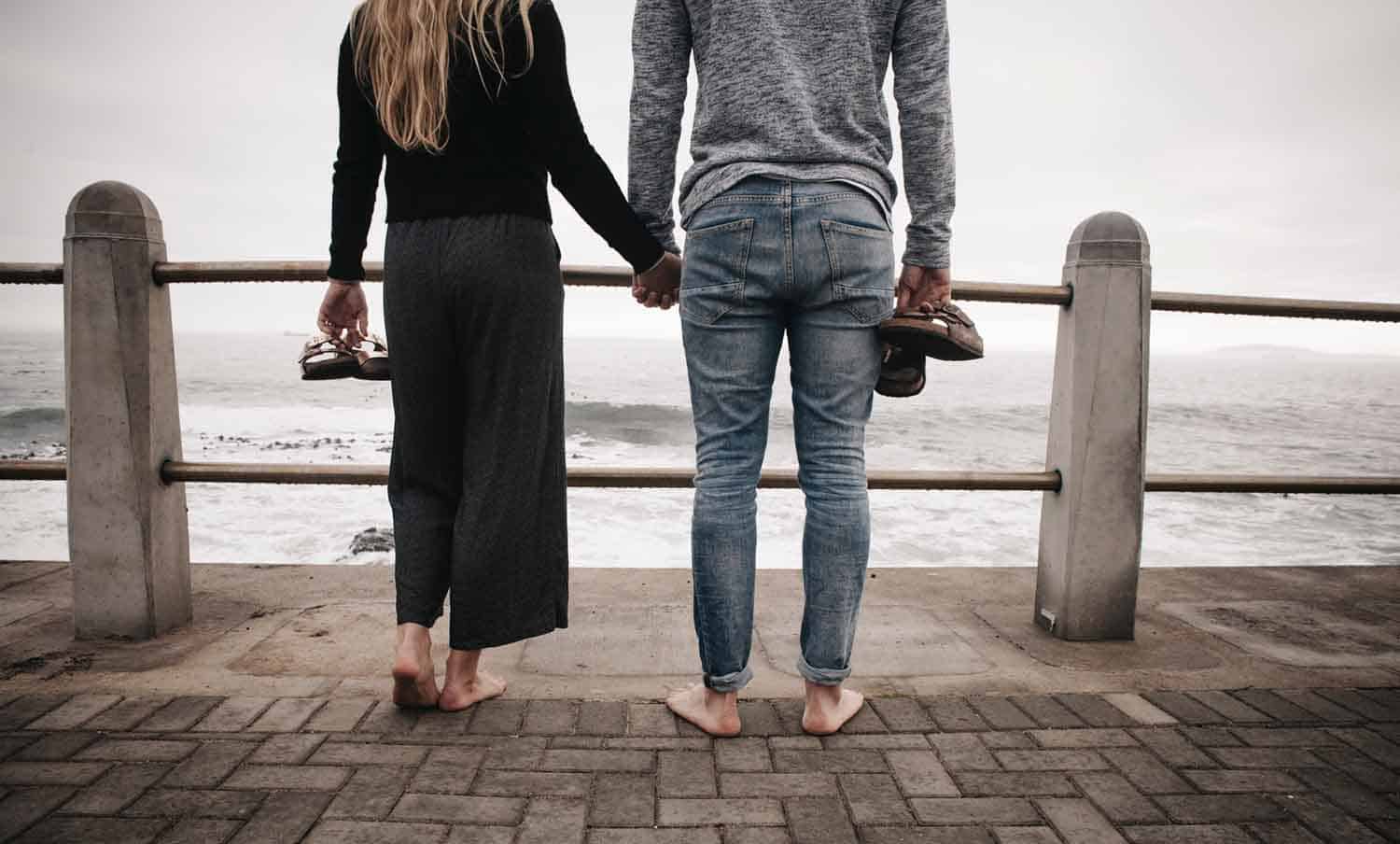 Coupole holding hands, standing barefoot in front of sea and holding Birkenstocks in their hands