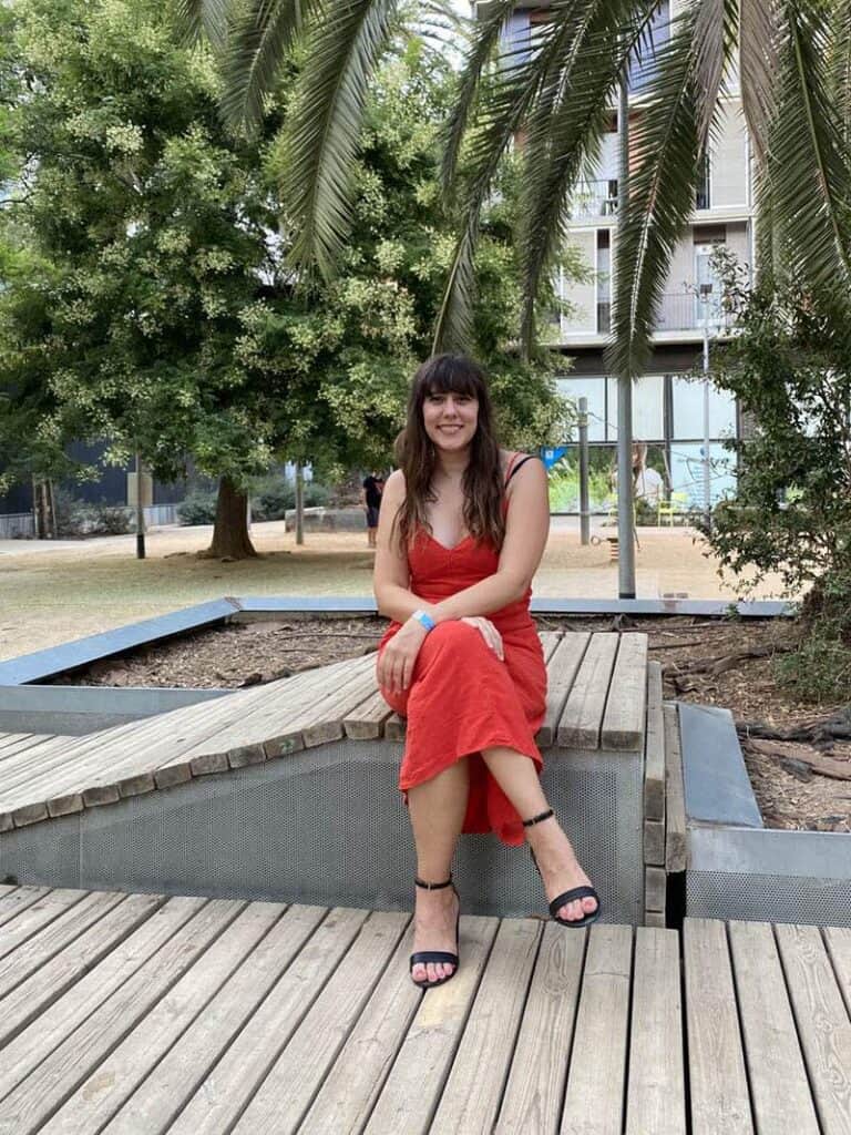 The author (Caitlin Galer-Unti) shown sitting in a park in Barcelona wearing red dress and Veerah Dian sandals